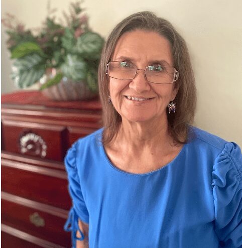 A woman in blue shirt and glasses standing next to a dresser.